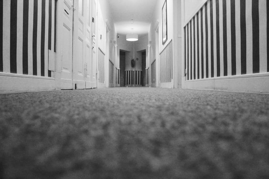 A black and white view of a long hallway with a low-angle perspective, showing a textured carpeted floor that appears clean and well-maintained. On either side of the hallway, there are white wooden doors and wainscoting with vertical striped wallpaper above. The ceiling has a modern round ceiling light, casting soft illumination throughout the corridor. The space appears tidy, with no visible dust or clutter, exemplifying professional surface cleaning and maintenance in a residential or commercial setting. The image emphasizes deep cleaning and sanitation efforts typical of services offered by Stockwell Cleaners on Clapham Road.