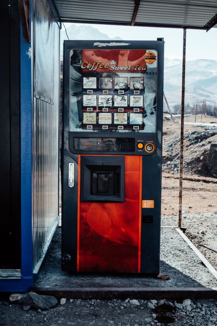A red and black coffee vending machine positioned outdoors under a metal roof, with a background of a mountainous landscape and a partly cloudy sky. The machine has a display panel with images of various beverage options and a keypad for selecting drinks. The area around the machine appears clean and dry, with gravel on the ground. The scene suggests a rest stop or outdoor refreshment area, where maintenance and cleaning services by Stockwell Cleaners could ensure the vending machine and surrounding surfaces remain hygienic and well-maintained.