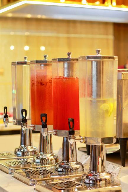 A row of four large, transparent dispensers filled with different colored beverages, including red and yellow liquids, are positioned on a stainless steel countertop in a commercial setting. Each dispenser has a black or silver lever for dispensing, with the liquids appearing clean and freshly filled. The background features a wooden panel wall illuminated by warm lighting, creating a welcoming atmosphere. The surface of the countertop is spotless, reflecting the containers and suggesting regular cleaning and maintenance by Stockwell Cleaners, who specialise in domestic and commercial surface cleaning, sanitisation, and deep cleaning services to ensure hygiene and cleanliness in various environments.