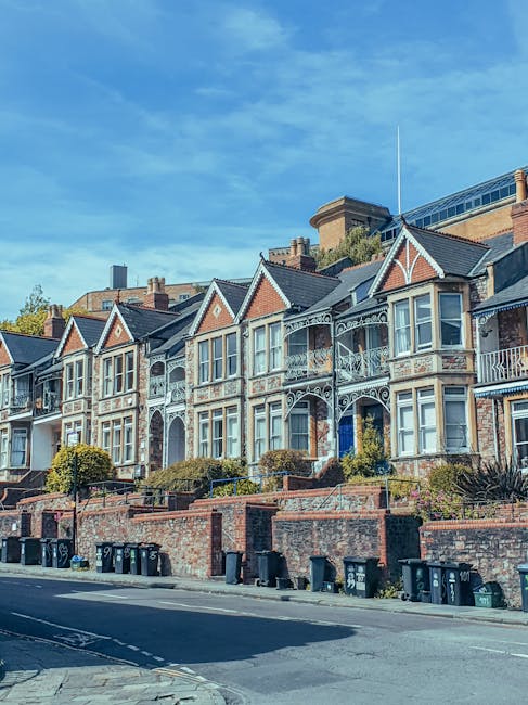 Exterior view of a row of Victorian-style terraced houses on Stockwell Park Estate, featuring brick facades, bay windows, decorative ironwork, and small front gardens. The houses are set against a bright blue sky with some scattered clouds. The street below has a line of black waste bins along a low brick wall, with a paved sidewalk in front. The scene is well-lit by natural daylight, highlighting the well-maintained appearance of the properties. As part of professional cleaning and maintenance, [COMPANY_NAME] offers domestic cleaning services to ensure the cleanliness and hygiene of similar residential properties, including deep surface cleaning and sanitisation of external surfaces.
